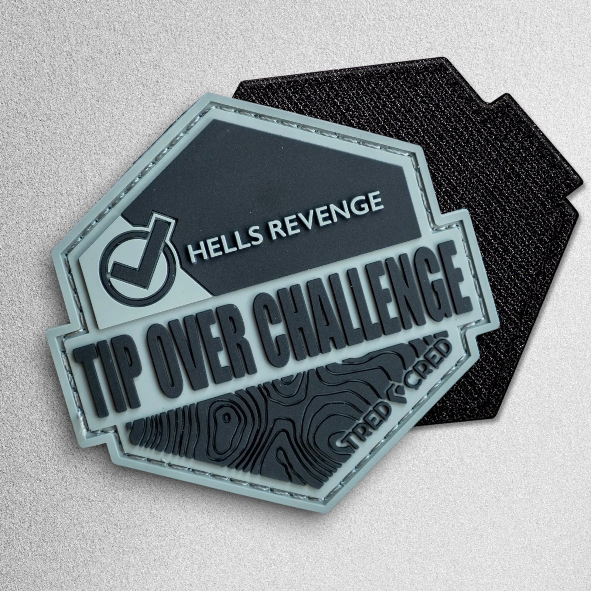 Tred Cred Tip Over Challenge hexagonal PVC patch in grey and black with topographic map detail and hook backing.