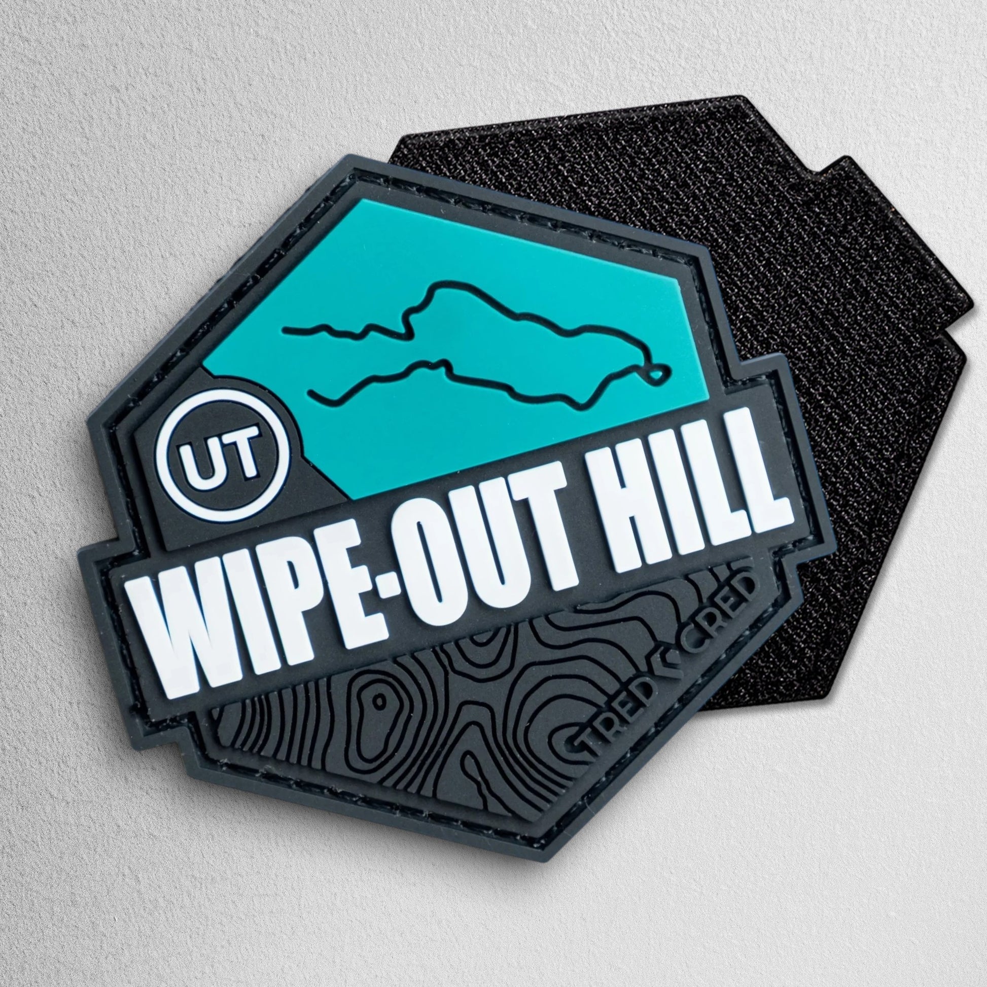 Tred Cred Wipe-Out Hill hexagonal PVC patch in teal and black with topographic map detail and hook and loop backing.